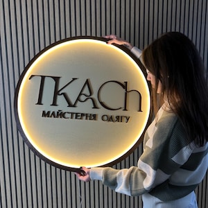 May include: A round wooden sign with the word "TKAC" in dark brown letters and the text "МАЙСТЕРНЯ ОДЯГУ" in a lighter brown. The sign has a warm, glowing light around the edge. The sign is being held up.