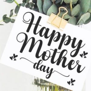 Puede incluir: Una tarjeta blanca con las palabras "Happy Mother's Day" en escritura negra est&aacute; sujeta por un clip dorado. Hojas y tallos de eucalipto verdes en el fondo, creando un arreglo floral natural.