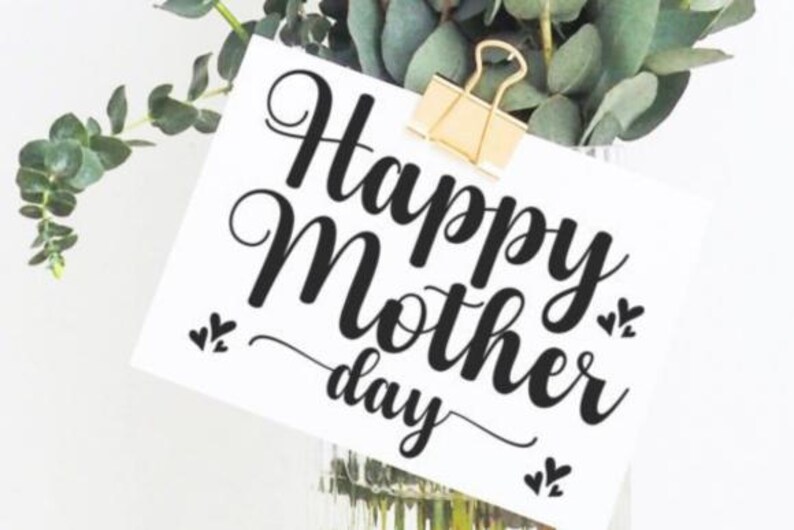 Puede incluir: Una tarjeta blanca con las palabras "Happy Mother's Day" en escritura negra est&aacute; sujeta por un clip dorado. Hojas y tallos de eucalipto verdes en el fondo, creando un arreglo floral natural.