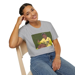 May include: A light grey t-shirt with a rectangular image of a person in a yellow uniform giving a thumbs-up gesture. The person is sitting on green grass. The t-shirt is being worn by a person.
