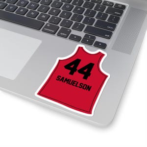 May include: A red sticker in the shape of a basketball jersey, with the number 44 in black and the name "SAMUELSON" below it. The sticker has a white border and is placed on a laptop.