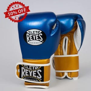 以下が含まれることがあります： 青とゴールドのボクシンググローブのペア。白いアクセント付き。グローブには「CLETO REYES」のロゴがあり、「HECHO EN MEXICO」と「PROFESSIONAL」と表示されています。赤と白の「50％OFF」ステッカーが左上にあります。