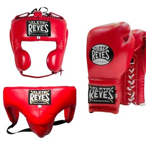 May include: Red boxing headgear, gloves, and groin protector set. The headgear has ear protection and a lace-up closure. The gloves are laced and the groin protector has a black strap. Each item has the "REYES" logo.