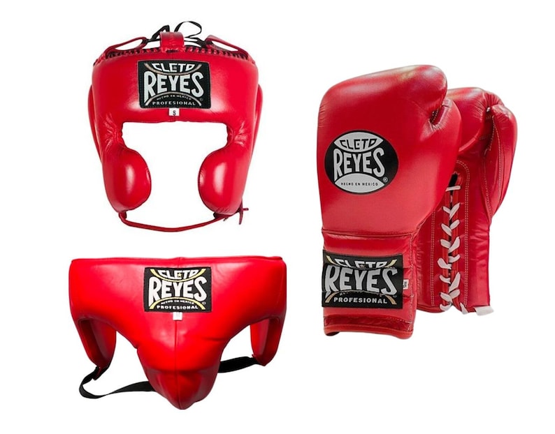 May include: Red boxing headgear, gloves, and groin protector set. The headgear has ear protection and a lace-up closure. The gloves are laced and the groin protector has a black strap. Each item has the "REYES" logo.