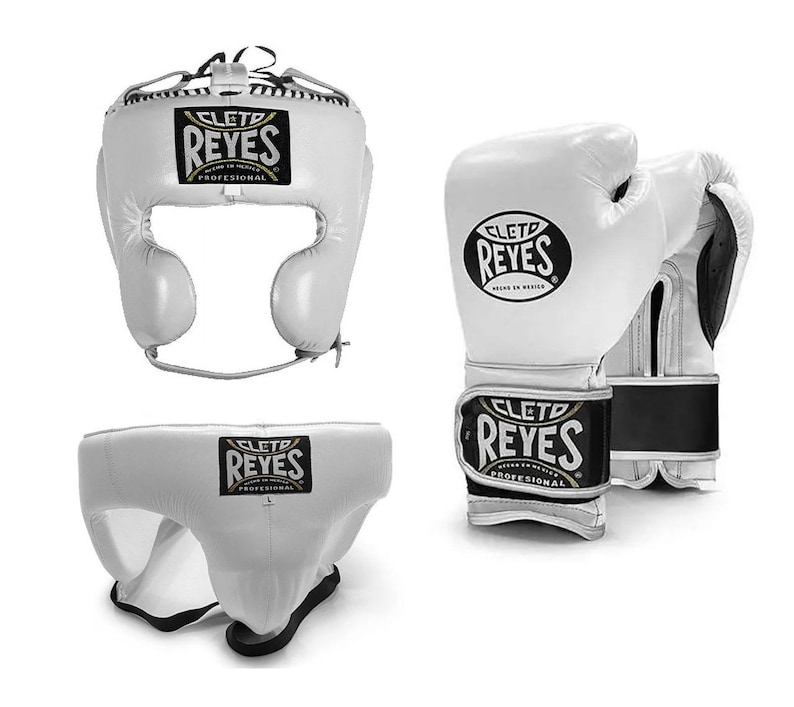 May include: White boxing headgear, gloves, and groin protector set. The headgear has a face opening and adjustable straps. The gloves have a wrist strap with the brand name "CLET0 REYES" in black and gold. The groin protector is white with a black and gold logo.