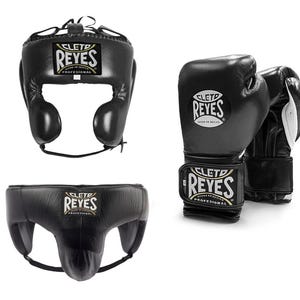 May include: Black boxing headgear, gloves, and groin protector set. The headgear has ear protection and a lace-up closure. The gloves and groin protector have the "REYES" logo. The gloves are black with white accents.
