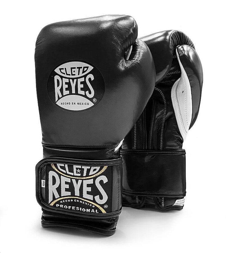 May include: Black leather boxing gloves with white accents. The gloves have the Cleto Reyes logo with the text "Cleto Reyes" and "Hecho en Mexico" on the front. The wrist strap has the text "Cleto Reyes" and "Profesional" on it.