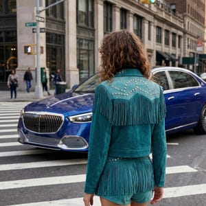 May include: Teal suede jacket and shorts set with fringe detailing and silver stud accents. The jacket has a decorative yoke and the shorts have a matching fringe hem. The outfit is worn in an urban setting with a blue car in the background.