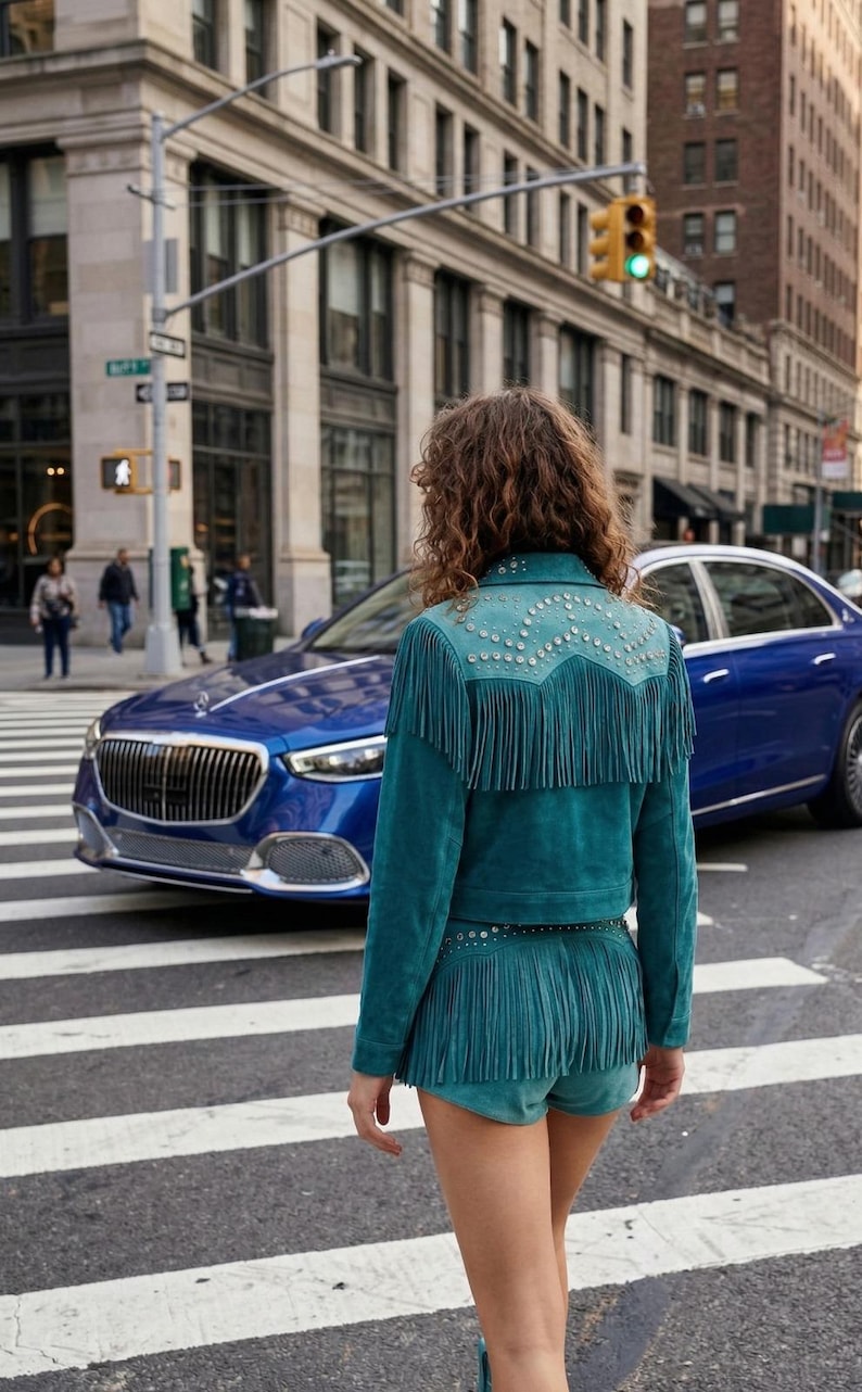 May include: Teal suede jacket and shorts set with fringe detailing and silver stud accents. The jacket has a decorative yoke and the shorts have a matching fringe hem. The outfit is worn in an urban setting with a blue car in the background.