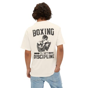 Puede incluir: Camiseta color crema con un gráfico negro de un boxeador y el texto "BOXING IS MY DISCIPLINE." El diseño presenta a un boxeador en posición de combate, con guantes de boxeo. La camiseta tiene cuello redondo y mangas cortas.