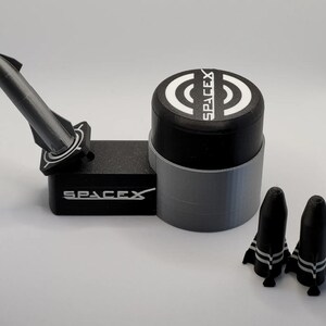 Può includere: Un modello di razzo SpaceX nero e grigio con una piattaforma di lancio e due modelli di razzo più piccoli. La piattaforma di lancio ha il logo SpaceX in bianco. Il modello di razzo è angolato verso l'alto e i modelli più piccoli sono in piedi.