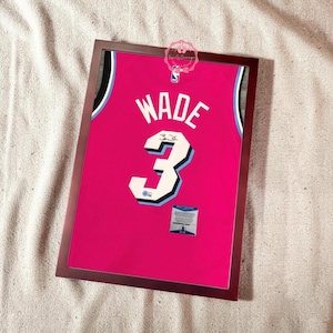 May include: Framed pink basketball jersey with the name "WADE" and the number "3" in white. The jersey has black and light blue accents and is displayed in a dark brown frame. A small label is visible at the bottom.
