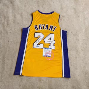 Może przedstawiać: Żółta koszulka do koszykówki z napisem "BRYANT" i numerem "24" w kolorze niebiesko-białym. Koszulka ma niebieskie akcenty po bokach i na dekolcie. Wydaje się być z autografem i zawiera metkę certyfikacyjną.