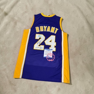 Może przedstawiać: Fioletowo-złoty koszulka do koszykówki z napisem "BRYANT" i numerem "24" w kolorze białym i złotym. Koszulka ma podpis i metkę certyfikacyjną. Koszulka to top bez rękawów.