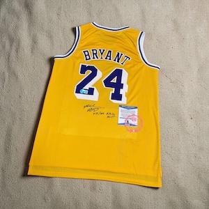 Może przedstawiać: Żółta koszulka do koszykówki z napisem "BRYANT" i numerem "24" w kolorze niebieskim. Koszulka ma białe wykończenia i podpis z przodu. Widoczna jest również mała naklejka autentyczności.