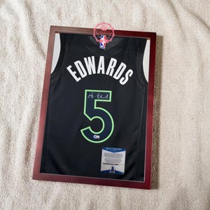 May include: Framed black basketball jersey with the name "EDWARDS" and the number "5" in white and green. The jersey is signed and includes a certificate of authenticity. The frame is a dark brown colour.