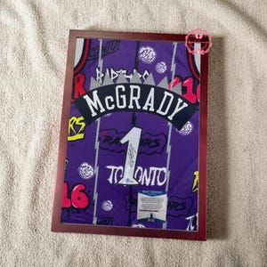 May include: Framed purple McGrady Toronto Raptors jersey with the number 1 and a signature. The jersey is in a brown frame. The jersey has white, red, and yellow accents. The jersey is on a light beige background.