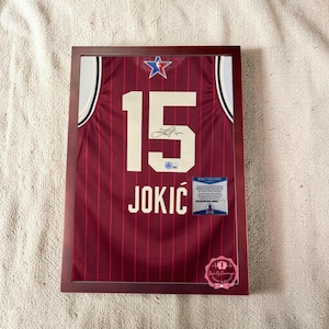 May include: Framed basketball jersey with the number 15 and the name JOKIC in white lettering. The jersey is maroon with white and blue accents and a star logo. Includes a certificate of authenticity and a signature.