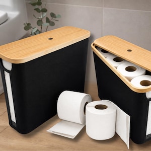 May include: Two black toilet paper storage containers with bamboo lids. One container is open, revealing rolls of white toilet paper. Two rolls of toilet paper are outside the containers. The containers are in a bathroom setting.
