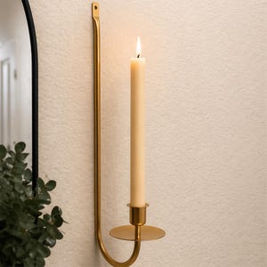 May include: A wall-mounted candle holder with a lit, cream-colored candle. The holder is made of gold-colored metal and has a curved design. The candle is tall and slender, with a small flame at the top. The holder is attached to a textured, off-white wall.