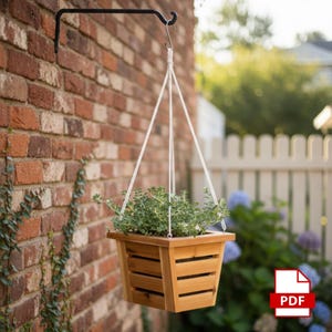 Peut inclure: Un pot de fleurs suspendu en bois rempli de plantes vertes. Le pot est suspendu par une corde blanche à un crochet en métal noir fixé à un mur de briques. Le pot a une forme carrée avec des lattes en bois horizontales.