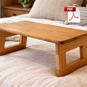 May include: A rectangular wooden bed tray with a natural finish. The tray has a flat surface and is supported by two sets of legs. The legs are constructed with a geometric design. The tray is placed on a bed with a white comforter.