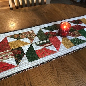 Könnte beinhalten: Gesteppter Tischläufer mit festlichem Design. Der Läufer hat ein Patchworkmuster aus roten, grünen, goldenen und floralen Stoffen. Eine rote Kerze steht auf dem Läufer. Der Läufer ist etwa 30 cm breit.