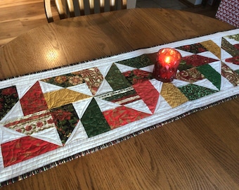 May include: A festive quilted table runner with a patchwork design. The runner features red, green, gold, and floral fabrics. A red candle sits on the runner. The runner is approximately 30 cm wide.
