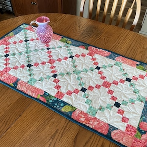 Può includere: Runner da tavolo trapuntato con un motivo floreale. Il runner presenta uno sfondo bianco con un motivo ripetuto di quadrati rossi, turchese e blu navy. I bordi sono bordati con una stampa floreale nei toni del rosa e del turchese. Una brocca di vetro rosa è sopra.