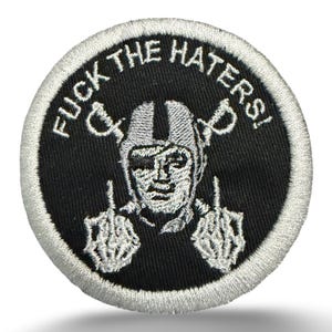 May include: A circular embroidered patch featuring a black background with a white border. The design depicts a figure wearing a helmet with two crossed swords behind it, and the text FUCK THE HATERS! arched above. The figure is giving the middle finger.