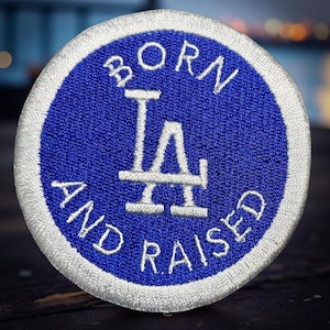 May include: Embroidered patch with a blue circle and white border. The patch features the text "BORN AND RAISED" in white, surrounding a white "LA" logo.
