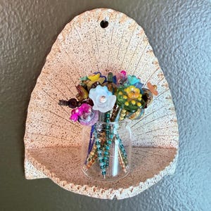 May include: A small glass vase filled with colourful beaded flowers. The vase sits on a speckled, off-white shelf attached to a wall. The shelf has a fan-like design. The flowers are various colours, including pink, yellow, and blue.