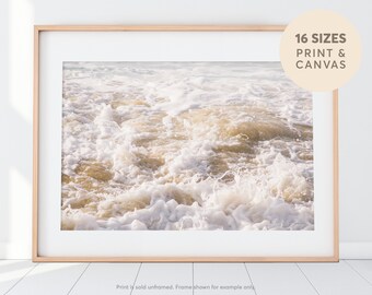 May include: A close-up photograph of foamy white ocean waves crashing on a sandy beach. The water is a light beige color and the foam is white. The image is taken from a low angle, giving the viewer a sense of being in the water.