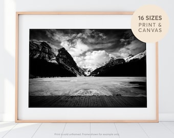 May include: Black and white photograph of a mountain range with a frozen lake in the foreground. A wooden dock extends into the lake. The sky is cloudy and dramatic.