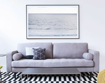 May include: A gray sofa with two pillows and a throw blanket sits in front of a framed photograph of a seascape. The sofa is in a living room with a black and white checkered rug and a coffee table.