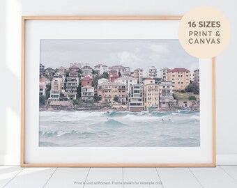 May include: A photograph of a colorful row of houses on a cliff overlooking a blue ocean with waves crashing on the shore. There are surfers in the water.