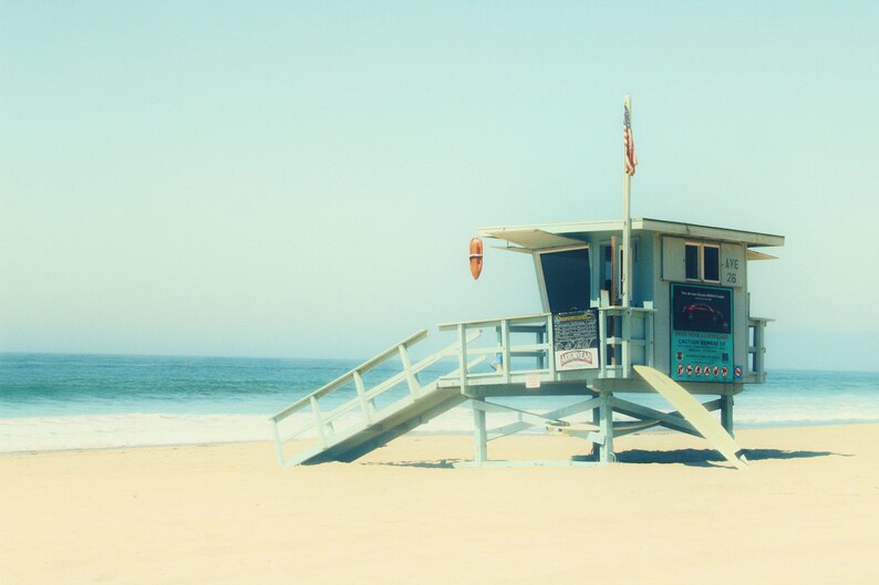 K&ouml;nnte beinhalten: Ein hellblauer Rettungsschwimmerstand mit einer Rampe und einer rot-wei&szlig;-blauen amerikanischen Flagge an einem Sandstrand mit blauem Wasser im Hintergrund.