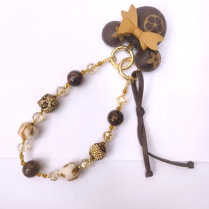 May include: A brown and gold beaded keychain with a leather bow and decorative accents. The keychain features a variety of beads, including round, faceted, and textured elements, connected by gold-toned chains and rings. A brown leather cord is attached.