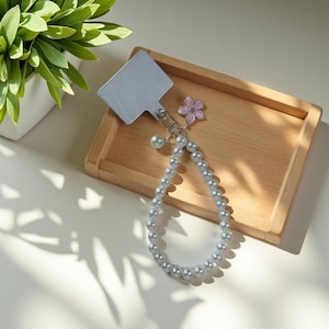 May include: A silver phone grip with a pearl beaded wrist strap. The strap is attached to a silver metal plate with a small pink flower charm and a pearl bead. The phone accessory is on a wooden tray.