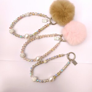 May include: Three beaded keychains with faux fur pom-poms. The keychains feature iridescent beads, faux pearls, and a small charm. One pom-pom is tan, and the other is pink. The keychains are on a white background.