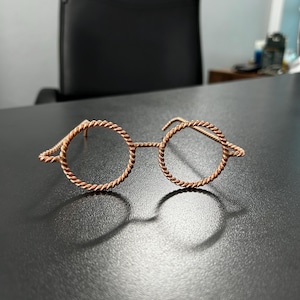 May include: A pair of round spectacles crafted from a single continuous piece of twisted copper-coloured wire. The frames feature a distinct rope-like texture throughout the circular rims and the bridge, with simple wire arms extending backwards.