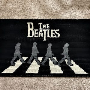 Könnte beinhalten: Schwarz-weißer Häkelteppich mit dem Text "The Beatles" und einer Silhouette der Bandmitglieder, die über einen Zebrastreifen gehen.