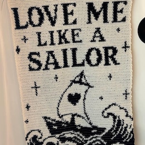 Könnte beinhalten: Ein handgefertigter, cremefarbener Wandbehang aus Häkelarbeit mit dem Text "LOVE ME LIKE A SAILOR" in Schwarz. Ein schwarzes Segelboot mit einem Herz auf dem Segel ist auf der unteren Hälfte abgebildet, über schwarzen Wellen. Der Wandbehang wird von einem Holzstab gehalten.
