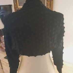May include: A black cropped bolero jacket featuring a textured, raised bubble pattern across the back and sleeves. The garment has long sleeves with fitted cuffs and a high collar, displayed on a mannequin to show the short, open-front design.