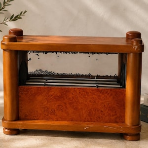 May include: A rectangular wooden rainmaker instrument featuring a polished light brown frame with a burl wood centre panel. The upper section contains a clear window filled with small dark beads, while the lower section houses internal metal rods.