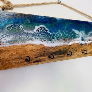 May include: A wooden key holder with a resin ocean wave design. The top features a blue and green ocean scene with white foamy waves. The bottom has a sandy beach appearance with six black hooks for keys. A rope is attached for hanging.