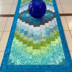 May include: A rectangular quilted table runner featuring a gradient of blue, teal, and lime green fabric squares with intricate swirling stitch patterns. A smooth, spherical, deep cobalt blue glass vase sits centered on the runner.