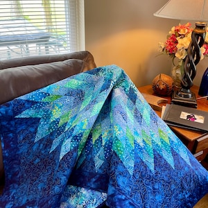 Könnte beinhalten: Eine handgefertigte Steppdecke in Blau- und Grüntönen. Die Steppdecke hat ein geometrisches Muster mit einem Farbverlauf, der von Dunkelblau zu helleren Blau- und Grüntönen übergeht. Die Steppdecke ist über ein braunes Ledersofa drapiert.