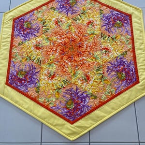 May include: Hexagonal quilted table runner with a floral pattern. The centre displays orange and yellow flowers, encircled by purple blooms. A red border and yellow edge complete the design.
