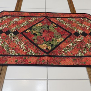 Puede incluir: Camino de mesa decorativo con un diseño floral de flor de Pascua. El camino de mesa tiene un fondo rojo con rayas diagonales de flores de Pascua rojas y blancas. Un borde negro enmarca los bordes, y un rombo central muestra un arreglo floral.